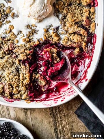 Apple blackberry crumble being scooped out of a white ceramic dish with a scoop of vanilla ice cream on top.