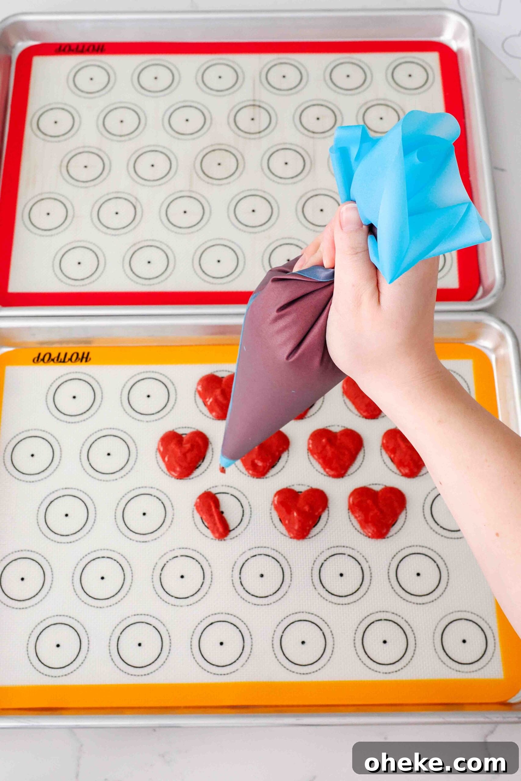Heart-Shaped Chocolate Raspberry Macarons Recipe 11 A hand pipes the left half of a heart-shaped macaron on silicone macaron mats.