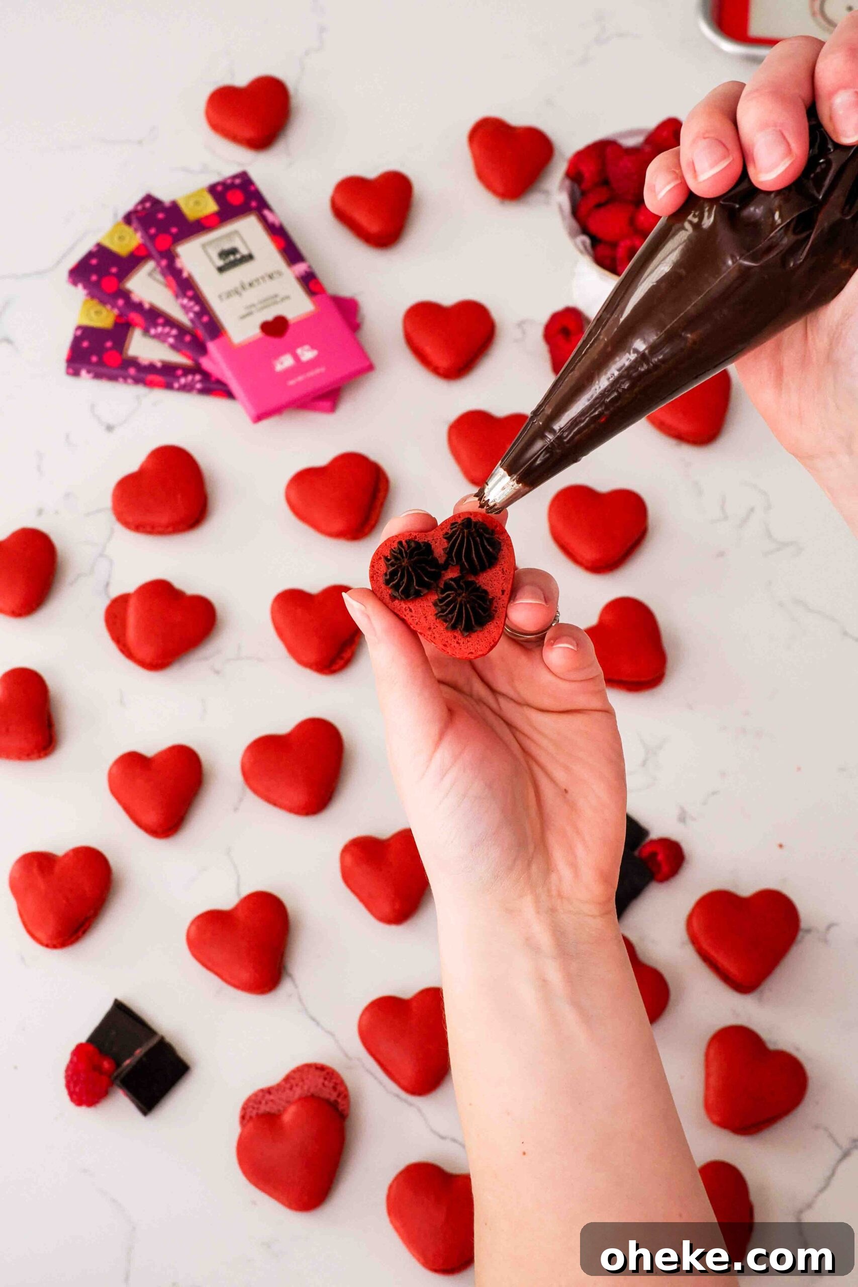 Heart-Shaped Chocolate Raspberry Macarons Recipe 17 A hand pipes three dots of raspberry chocolate ganache onto the underside of a heart-shaped macaron shell.