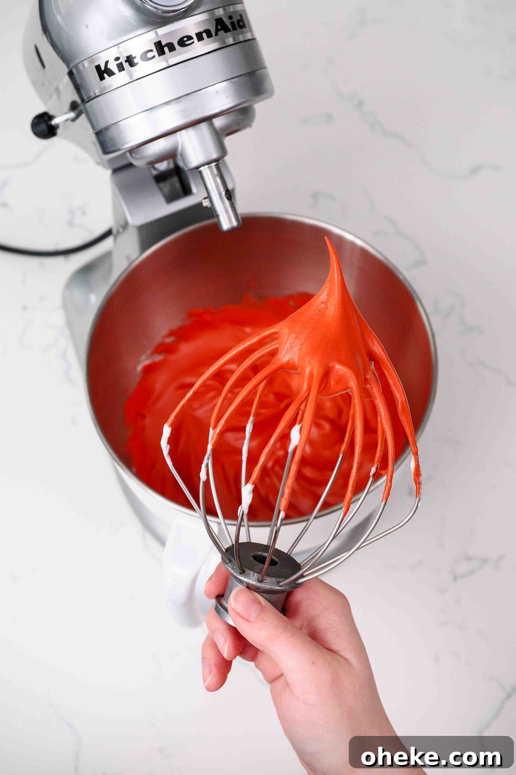 Heart-Shaped Chocolate Raspberry Macarons Recipe 7 A hand holds up a whisk with a stiff peak of red meringue pointing up.