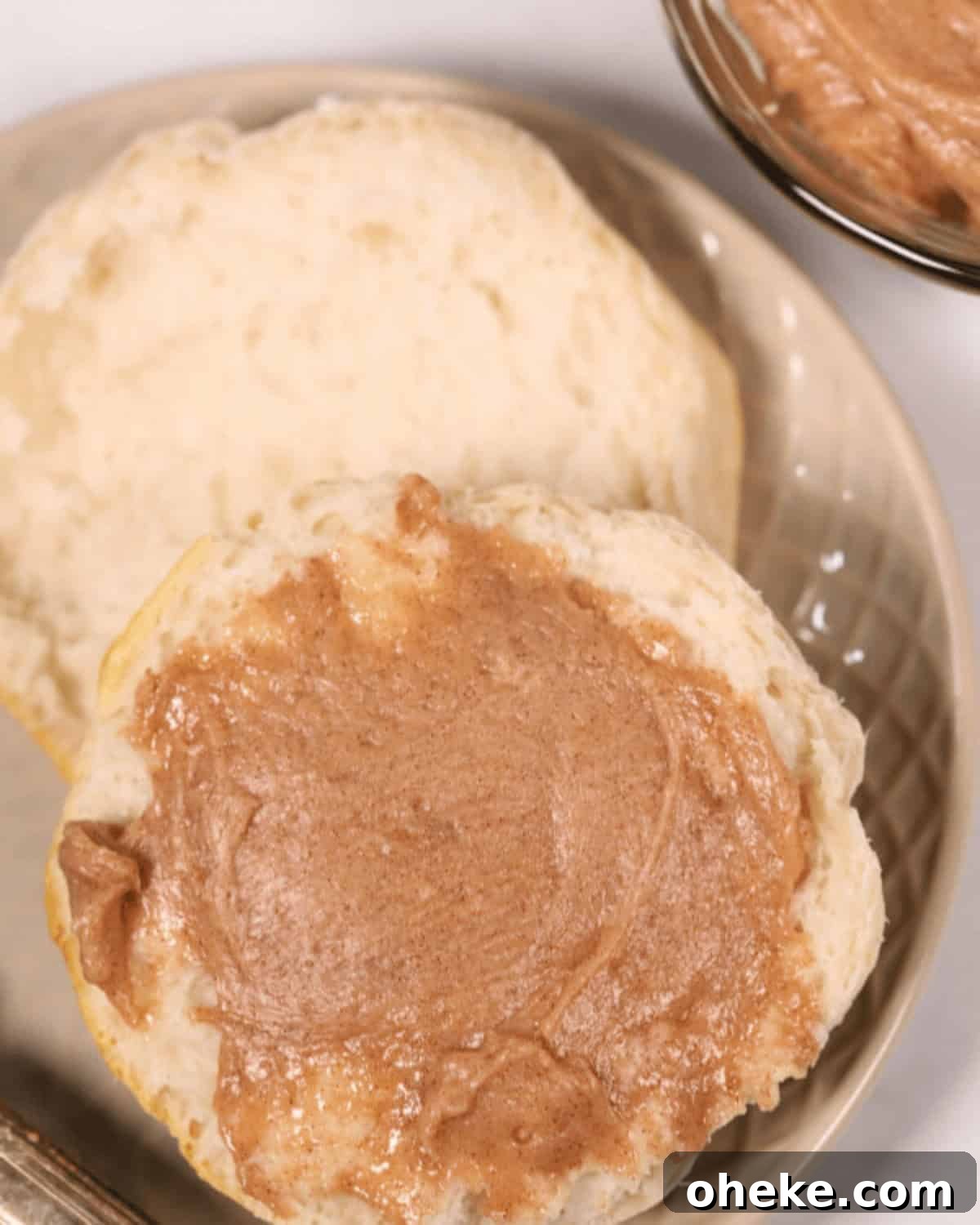 A biscuit, split in half and one side spread with Texas Roadhouse Butter, is placed on a beige plate. Nearby is a bowl containing more delightful cinnamon butter.