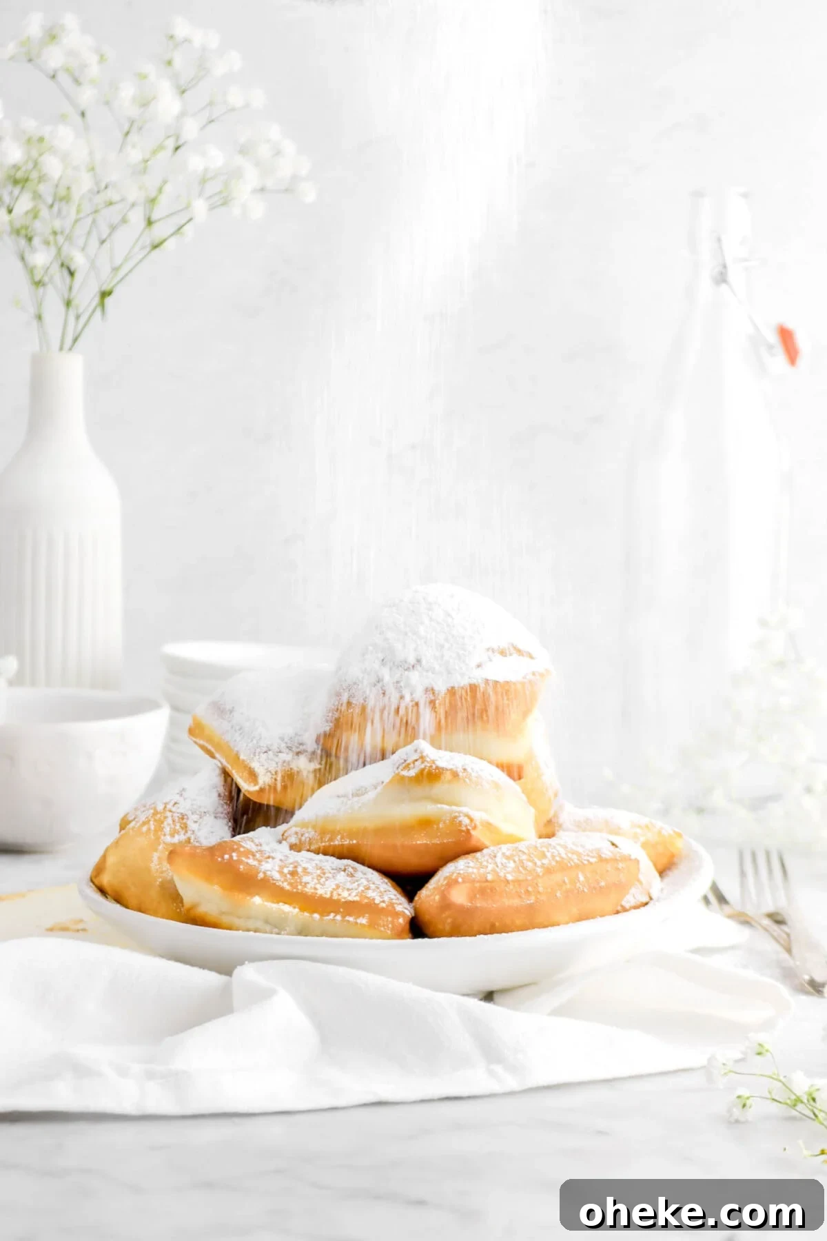 close up of stacked beignets being dusted with powdered sugar in a white bowl