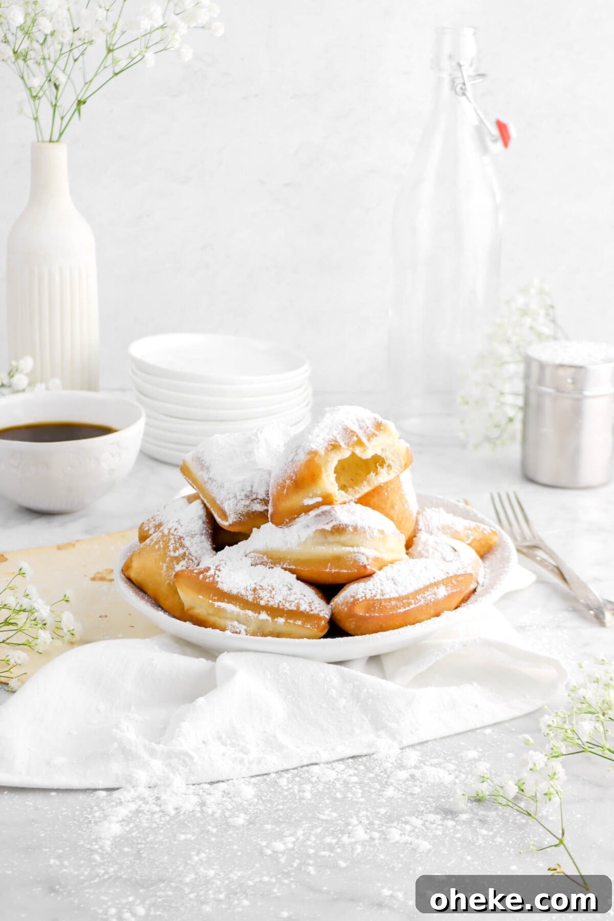 angled photo of beignets in white bowl with bite missing from top beignet on white napkin with old book pages, two forks, and stack of white plates behind