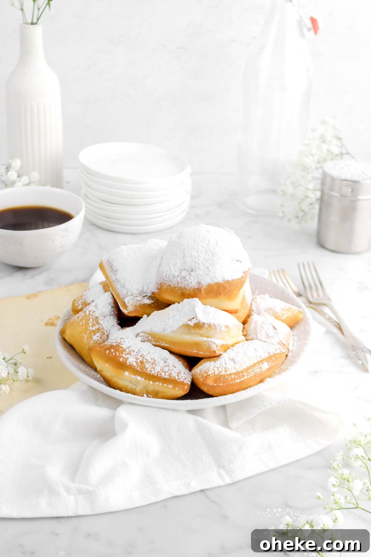 angled photo of beignets in white bowl withs tack f plates, flowers, cup of coffee, forks, and white napkin