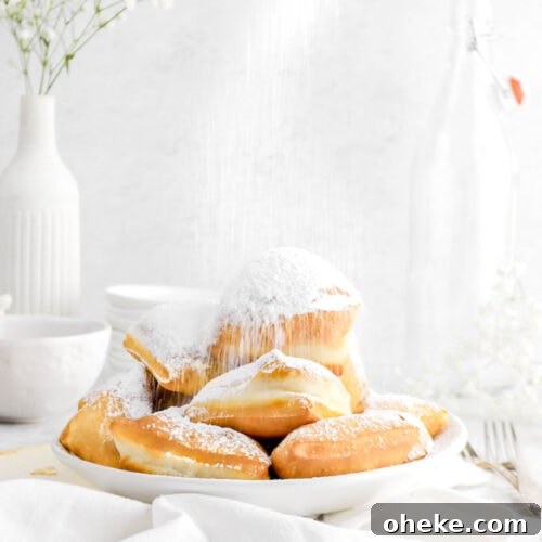 close up of stacked beignets being dusted with powdered sugar in a white bowl