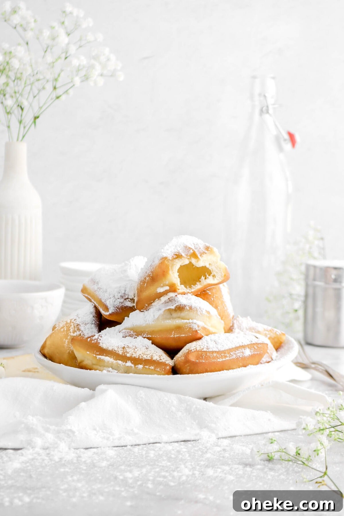 front shot of stack of beignets in white bowl with bite taken from the top one with flowers and white napkin