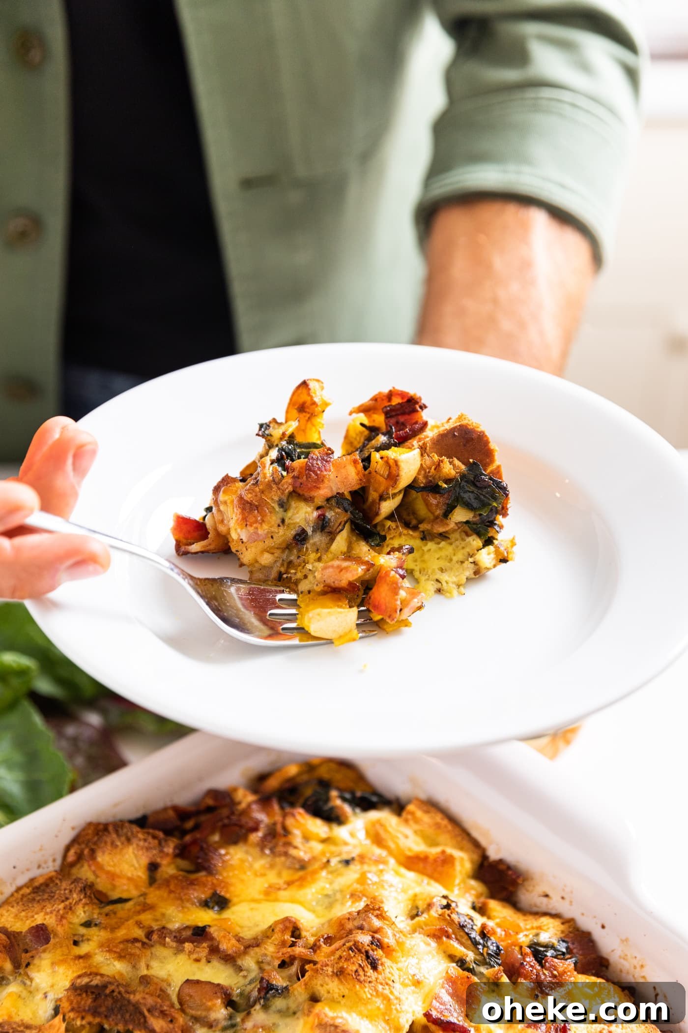 Hand holding white plate filled with scoop or squash strata with fork taking out a piece with rest of strata in white casserole dish below
