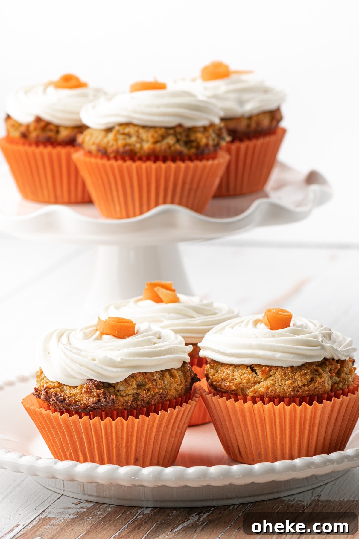 White plate with three muffins in the foreground and a stack of carrot cake muffins on a white ruffled cake stand in the back 