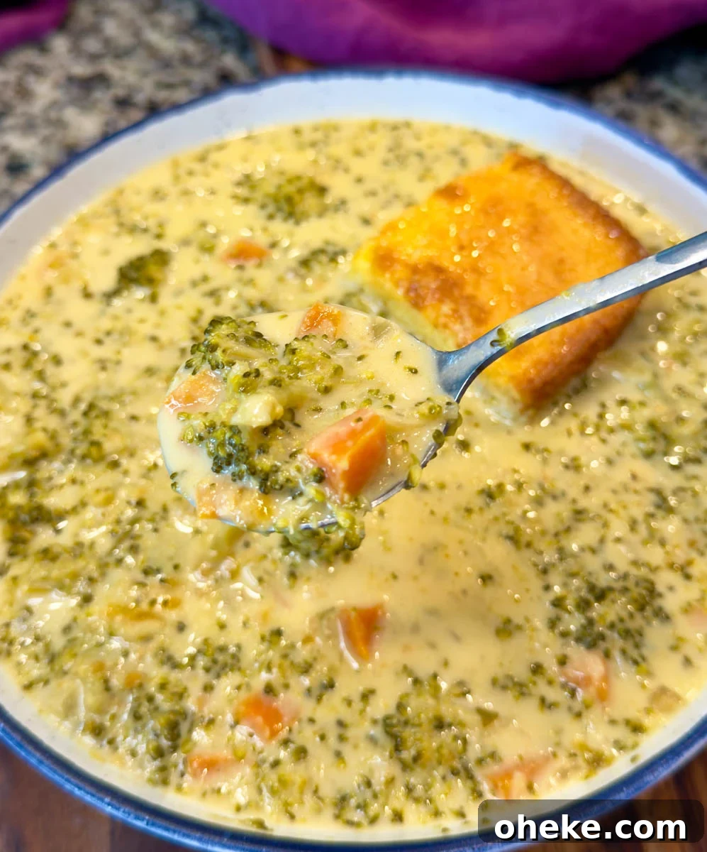 Slow Cooker Broccoli Cheddar Soup (Crockpot Recipe) 6 Close-up of a spoonful of broccoli cheddar soup lifted from a bowl, showing tender broccoli and carrots in creamy cheese broth with a square of cornbread in the background.