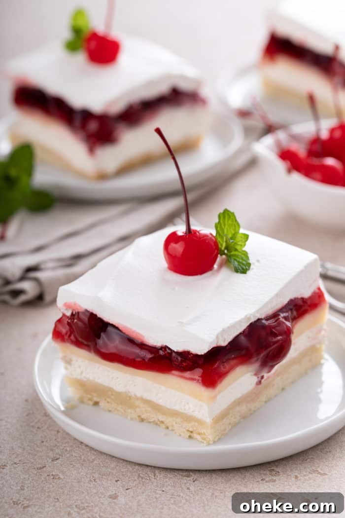 White plate with a slice of layered cherry cheesecake dessert topped with a maraschino cherry and sprig of mint.