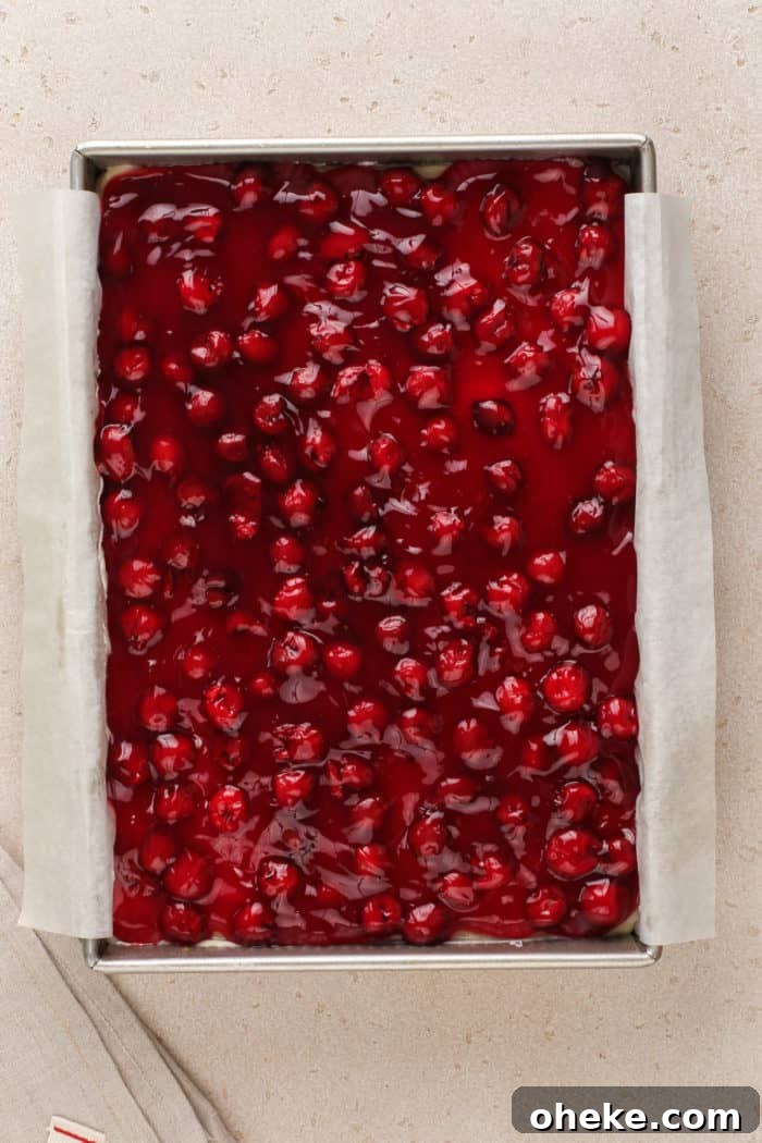 Cherry pie filling spread onto layered cherry cheesecake dessert in a baking pan.