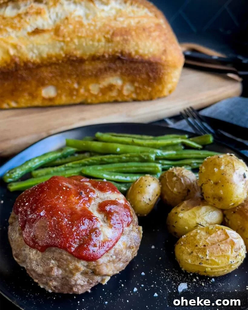 Mini meatloaf, roasted potatoes and green beans on a dark plate with a loaf of sourdough bread behind it.