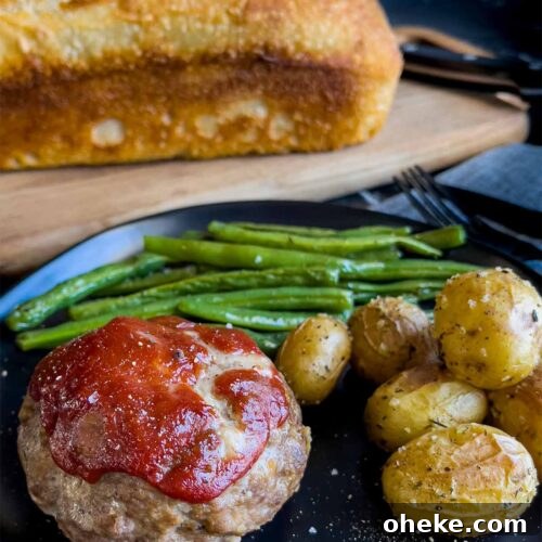 Mini meatloaf, roasted potatoes and green beans on a dark plate with a loaf of sourdough bread behind it.