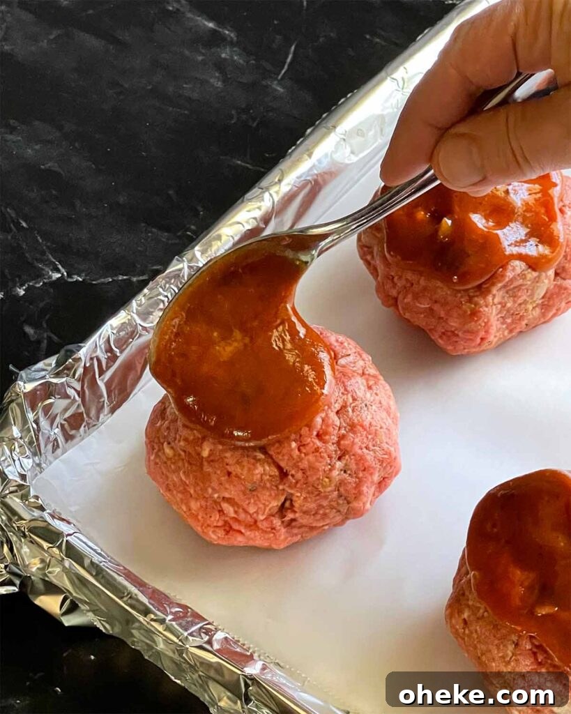 Unbaked mini meatloaves on a foil lined baking sheet with the sauce being spooned on top of them.