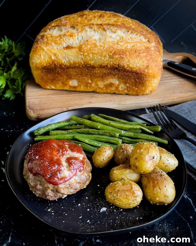 Mini meatloaf, roasted potatoes and green beans on a dark plate with a loaf of sourdough bread behind it.