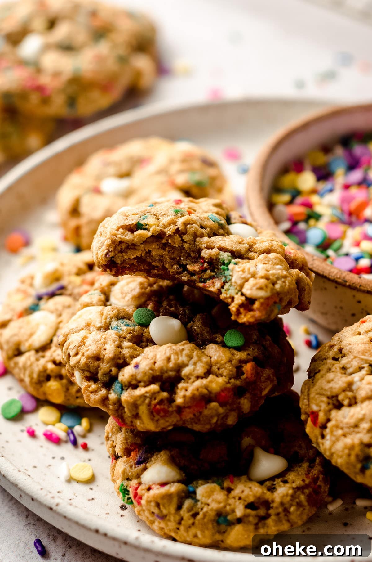 Funfetti White Chocolate Oatmeal Cookies Recipe 9 funfetti oatmeal cookies on a plate with a bowl of sprinkles and one cookie has a bite taken out of it