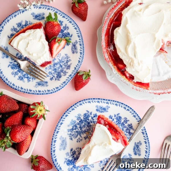 Overhead view of two plates with slices of strawberry pie topped with whipped cream, a whole pie, fresh strawberries, and white flowers on a pink surface.