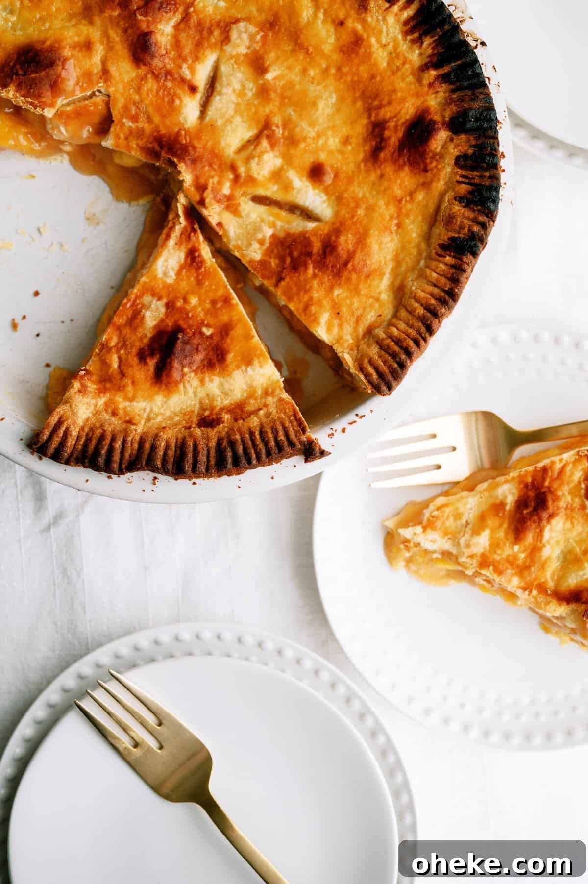 A golden-brown pie with a slice removed sits on a plate; a slice of the pie is served on a white plate beside gold forks on a white tablecloth.