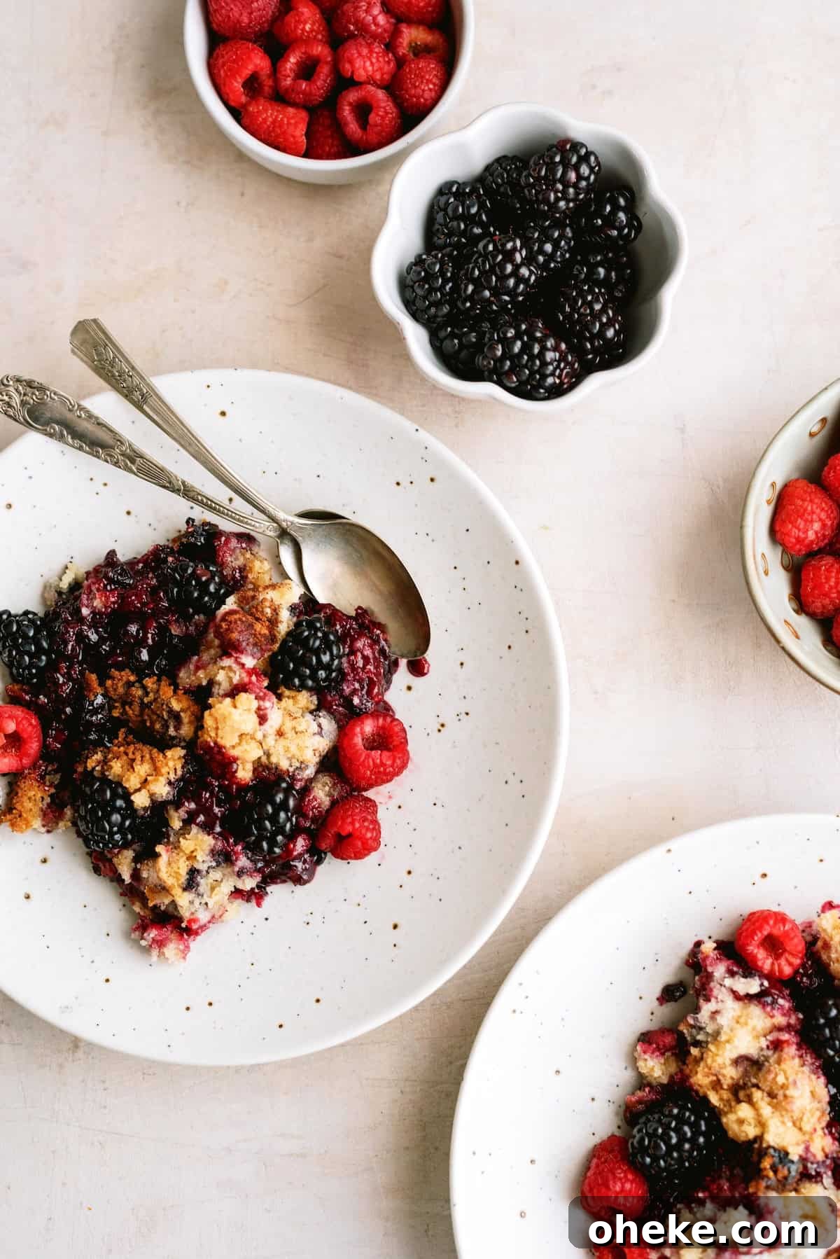 Plates of berry crumble with fresh raspberries and blackberries, served with spoons, beside small bowls of berries on a light surface.