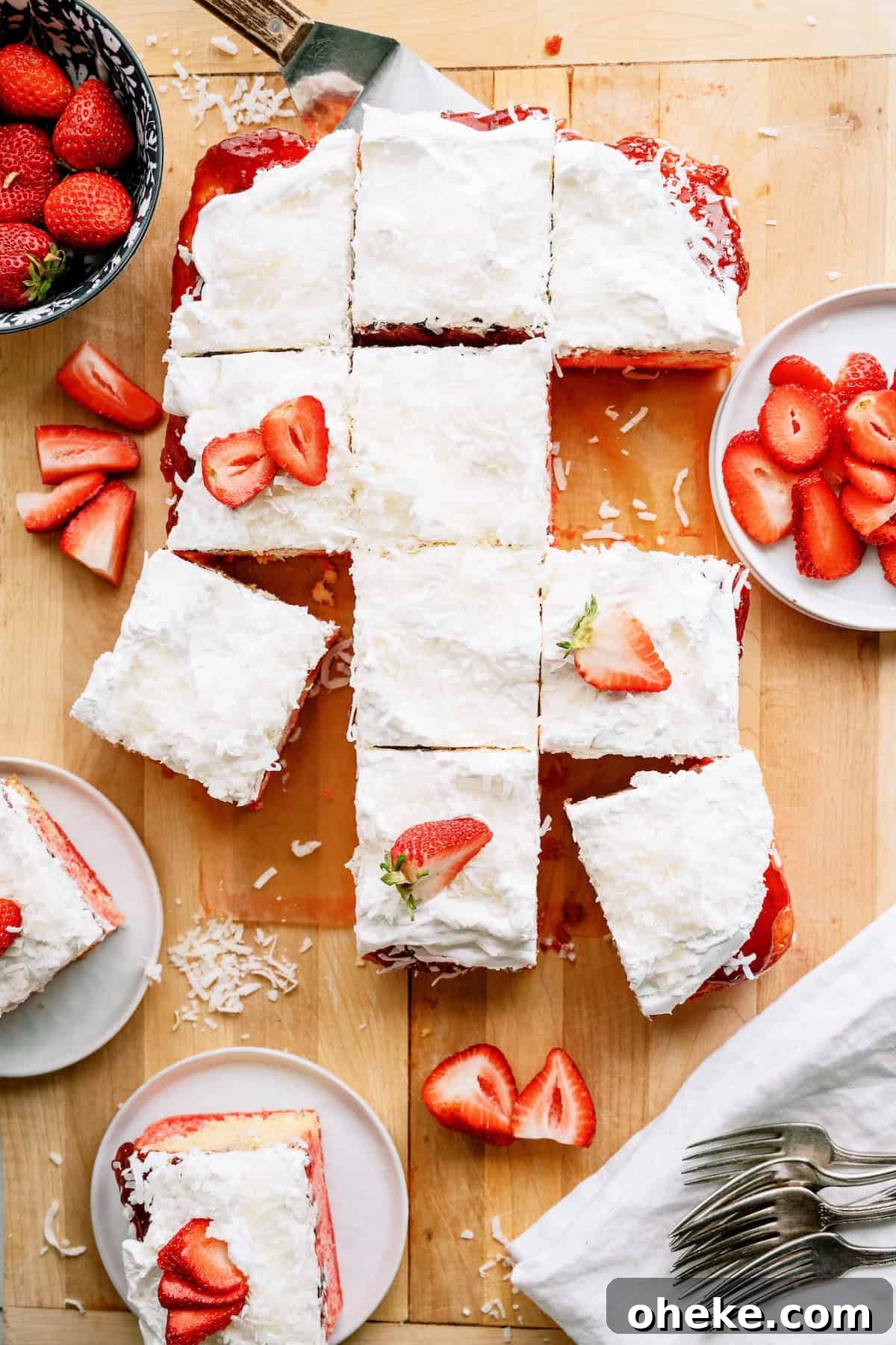 A rectangular coconut-topped cake is cut into squares, some garnished with sliced strawberries, and served on plates with extra strawberries on the side.