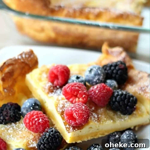Two squares of German Pancakes on a plate finished with powdered sugar with berries. Pan of remaining German Pancakes in the background.