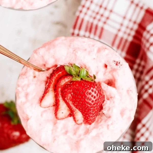 A glass bowl filled with pink strawberry mousse, topped with sliced fresh strawberries, sits on a white surface near a red and white plaid napkin.