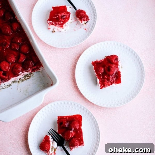 Three white plates with slices of raspberry dessert topped with red jelly, next to a baking dish containing the remaining dessert. Two plates have black forks.