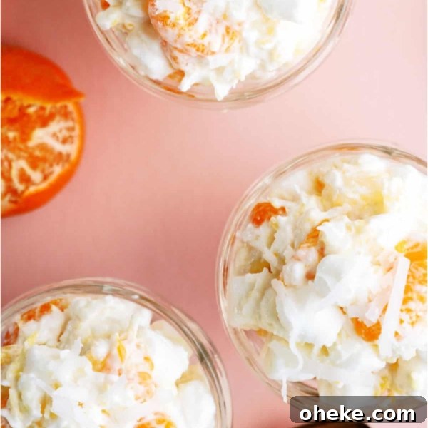 Three glass bowls filled with a creamy fruit salad containing orange segments, pineapple pieces, shredded coconut, and mini marshmallows, placed on a pink surface with spoons.