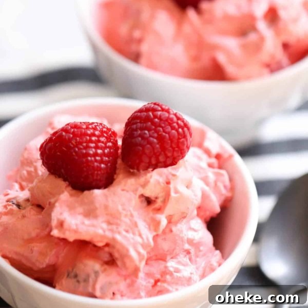 Two white bowls filled with pink creamy dessert topped with fresh raspberries, placed on a striped cloth next to a spoon.
