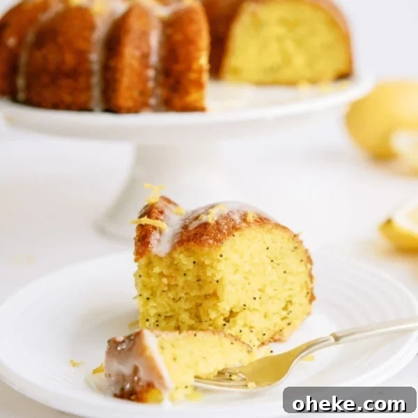 A slice of Lemon Poppy Seed Bundt Cake on a white plate with the remaining bundt cake on a cake stand in the background.