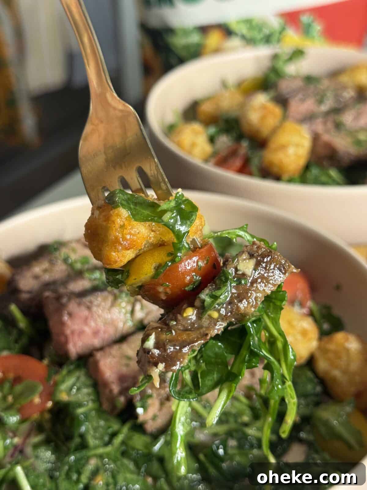 Steak and Potato Salad with Garlic Herb Dressing 3 A fork holds a bite of salad with arugula, cherry tomato, steak, and a crispy tater tot over a bowl filled with more steak, greens, and tater tots.