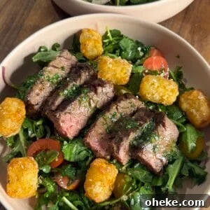 A bowl filled with sliced steak on a bed of mixed greens, cherry tomatoes, red onions, and topped with golden tater tots, all set on a wooden table.