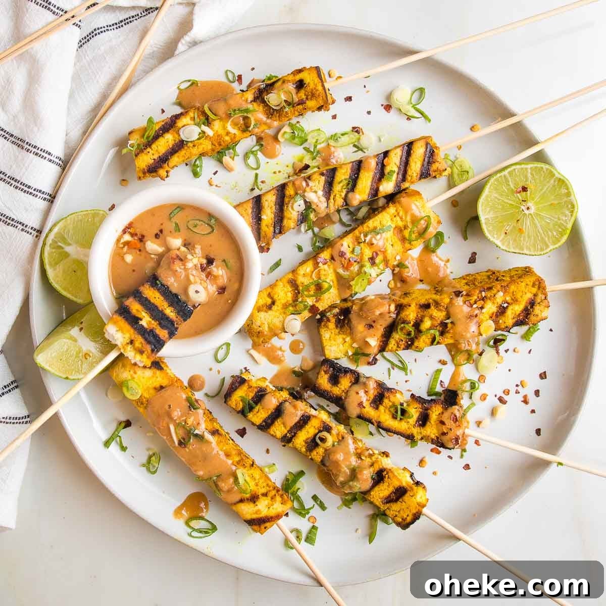 Round plate with grilled tofu satay skewers. One skewer with grill marks dipping into a small dish of peanut sauce.