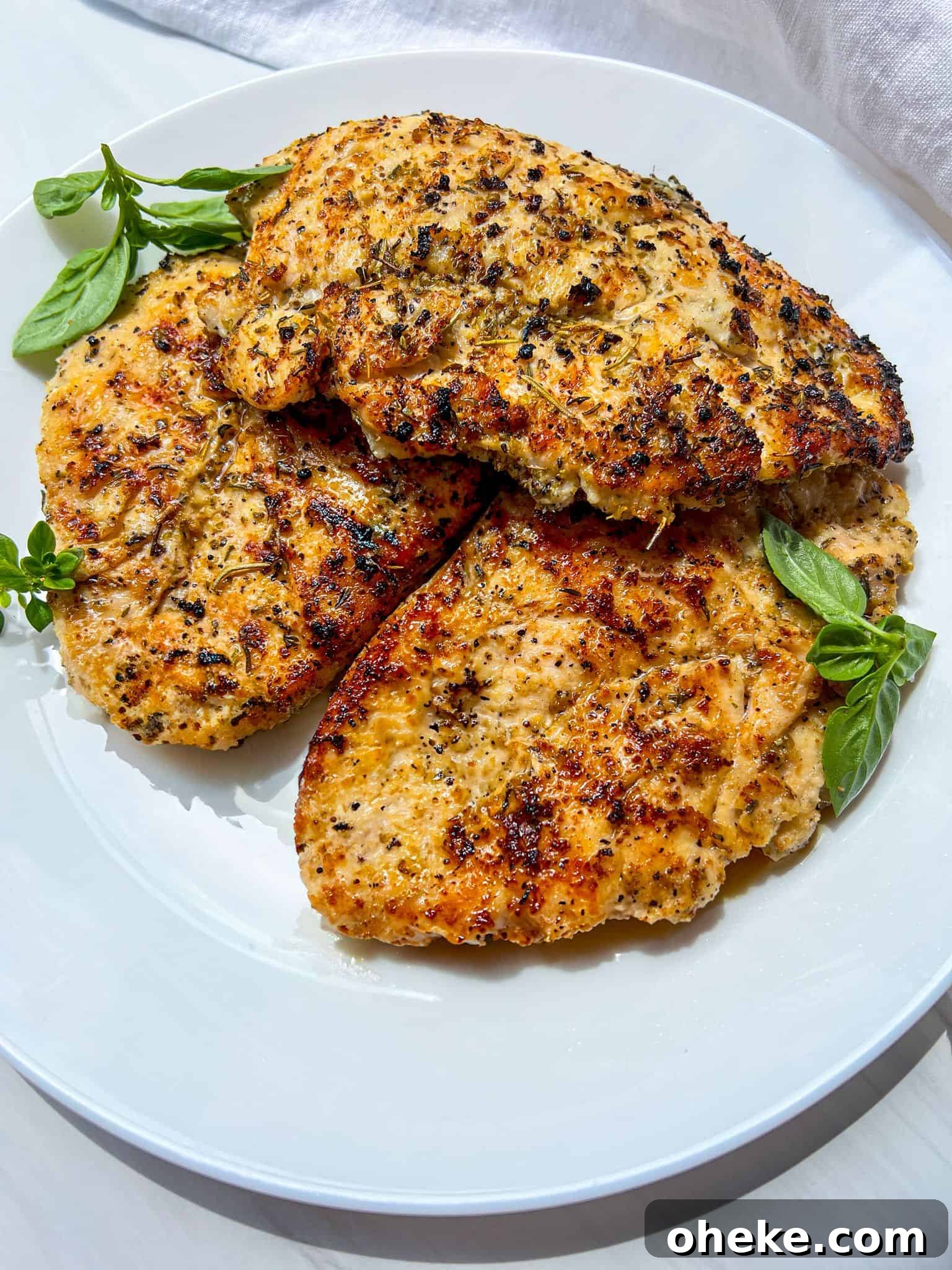 Three pieces of golden-brown, seasoned grilled chicken breast garnished with fresh basil leaves on a white plate.