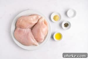 A white plate with two raw chicken breasts is placed on a marble surface. Nearby are small bowls containing olive oil, black pepper, salt, and a yellow seasoning blend.