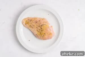 A raw chicken breast seasoned with black pepper and spices sits on a white plate against a light marble background.