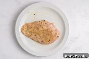 A raw, seasoned chicken breast sits on a white plate. The chicken is sprinkled with black pepper and other spices, and the plate rests on a light-colored marble surface.