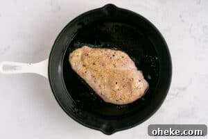 A raw, seasoned chicken breast is cooking in oil in a black skillet with a white handle, set on a light marble surface.