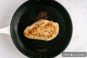 A seasoned, cooked chicken breast sits in a black skillet with oil, viewed from above on a light marble surface.