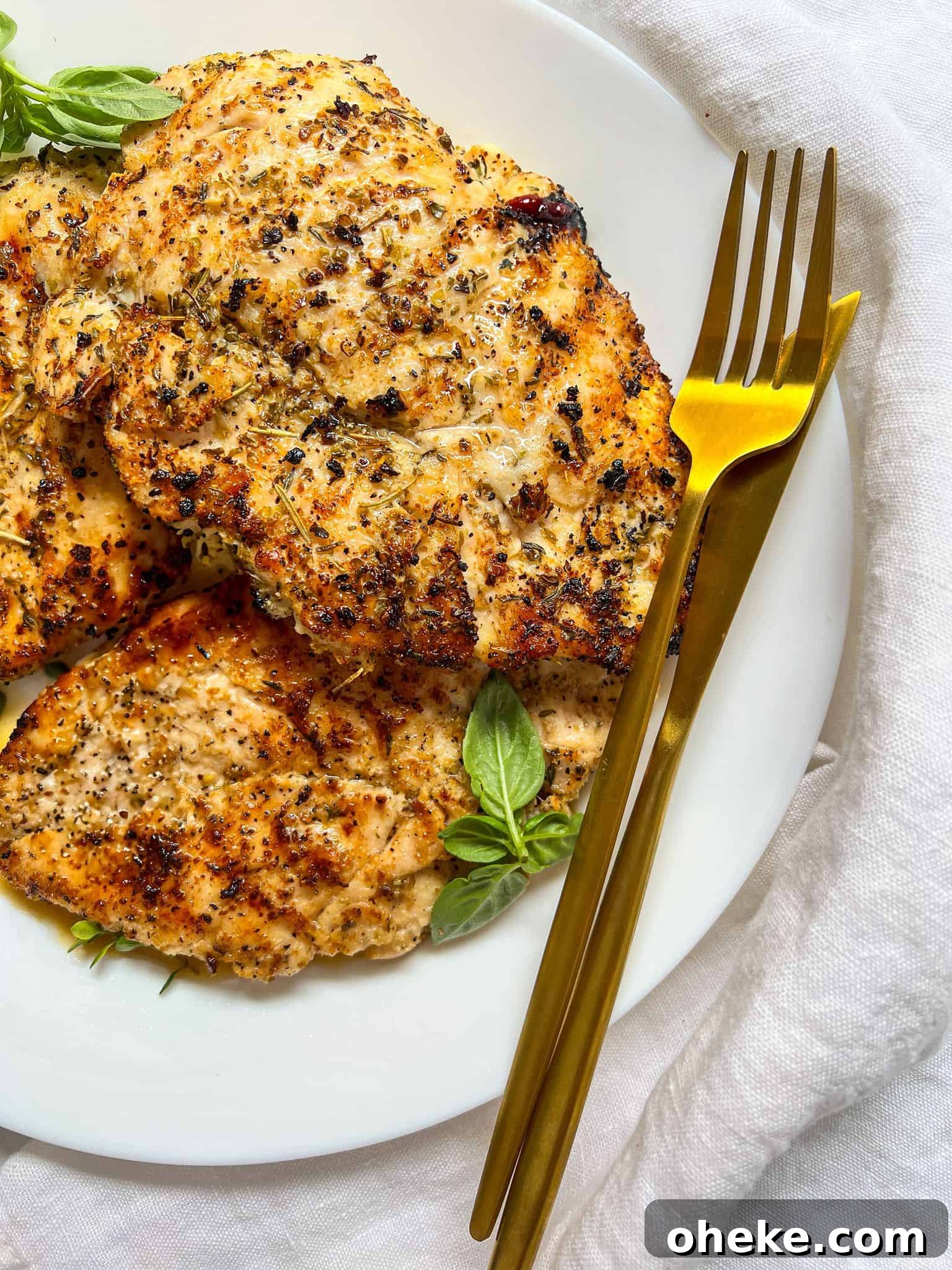 Grilled, seasoned chicken breasts on a white plate garnished with fresh basil, next to a gold fork and knife, set on a white cloth background.