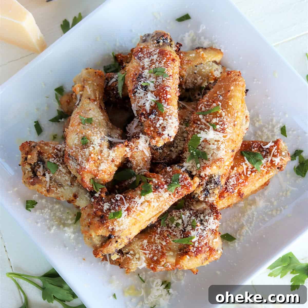 Square image of chicken wings piled high on a white plate and sprinkled with fresh parmesan and parsley
