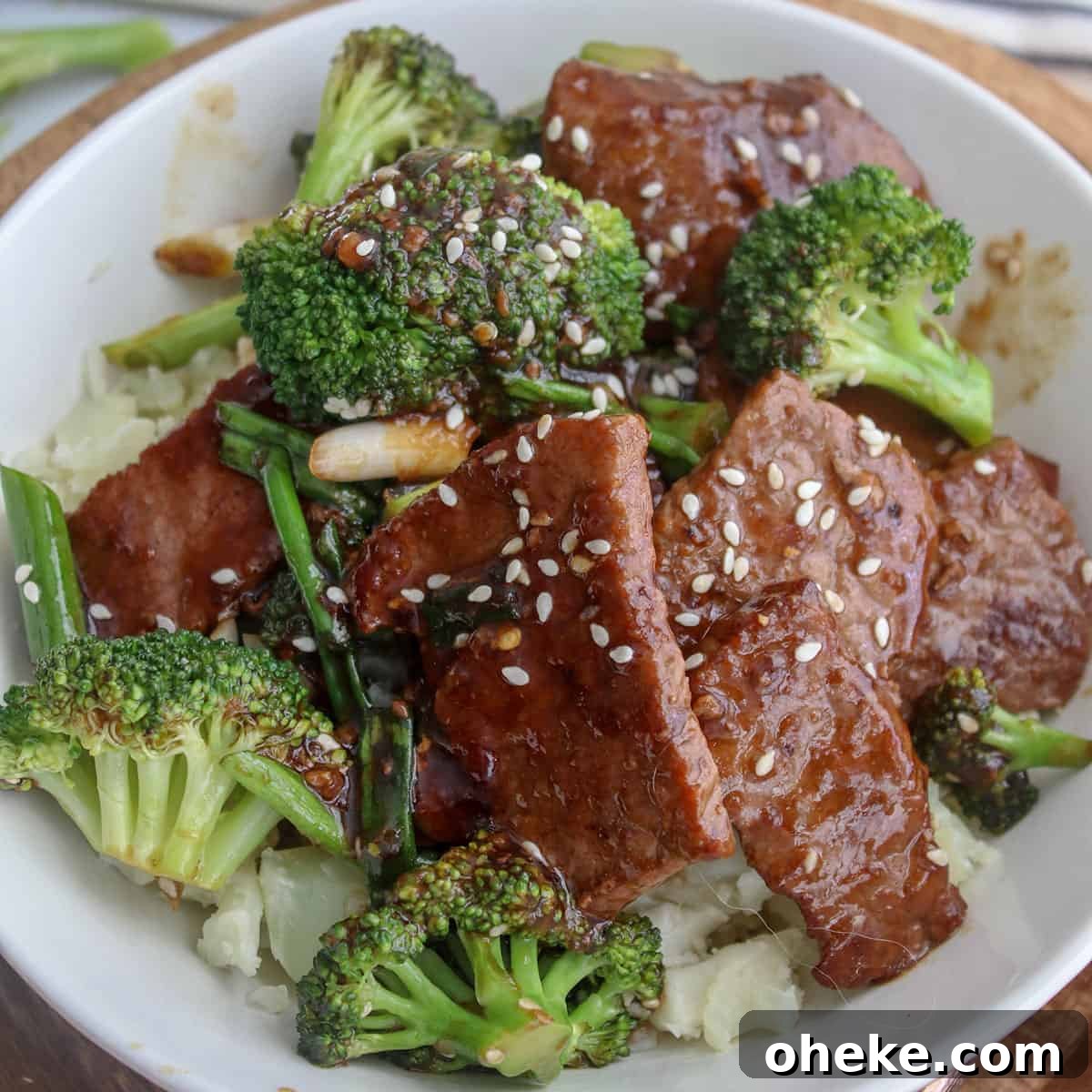square closeup image of mongolian beef and broccoli served in a white bowl and garnished with sesame seeds.
