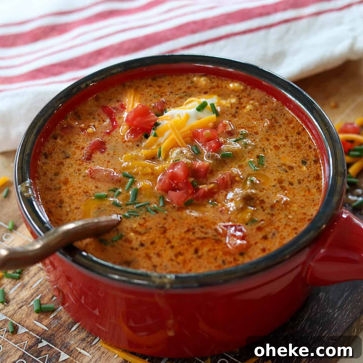 Square side image of soup served in a red bowl on a wooden cutting board. Soup is garnished with sour cream, tomato, green onion, and cheddar cheese.