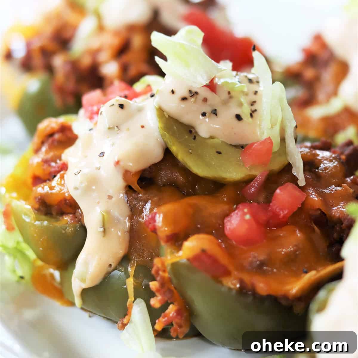 Square image closeup of cheeseburger stuffed peppers on a white plate, topped with lettuce, tomato, pickle.