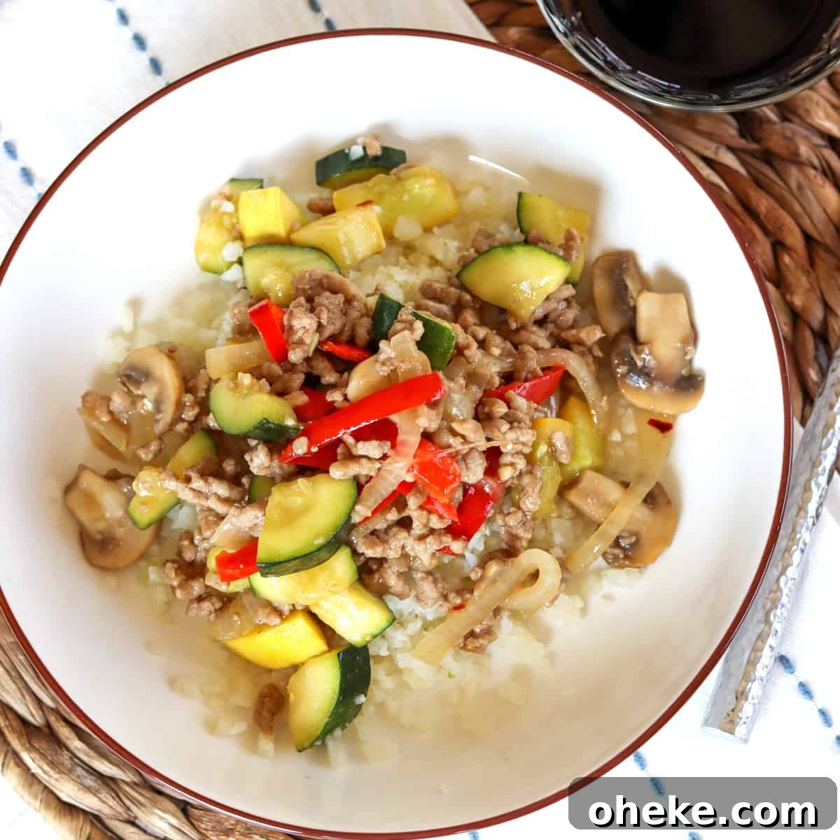 Square image of a bowl of pork stir fry served over rice with a side of stir fry sauce.
