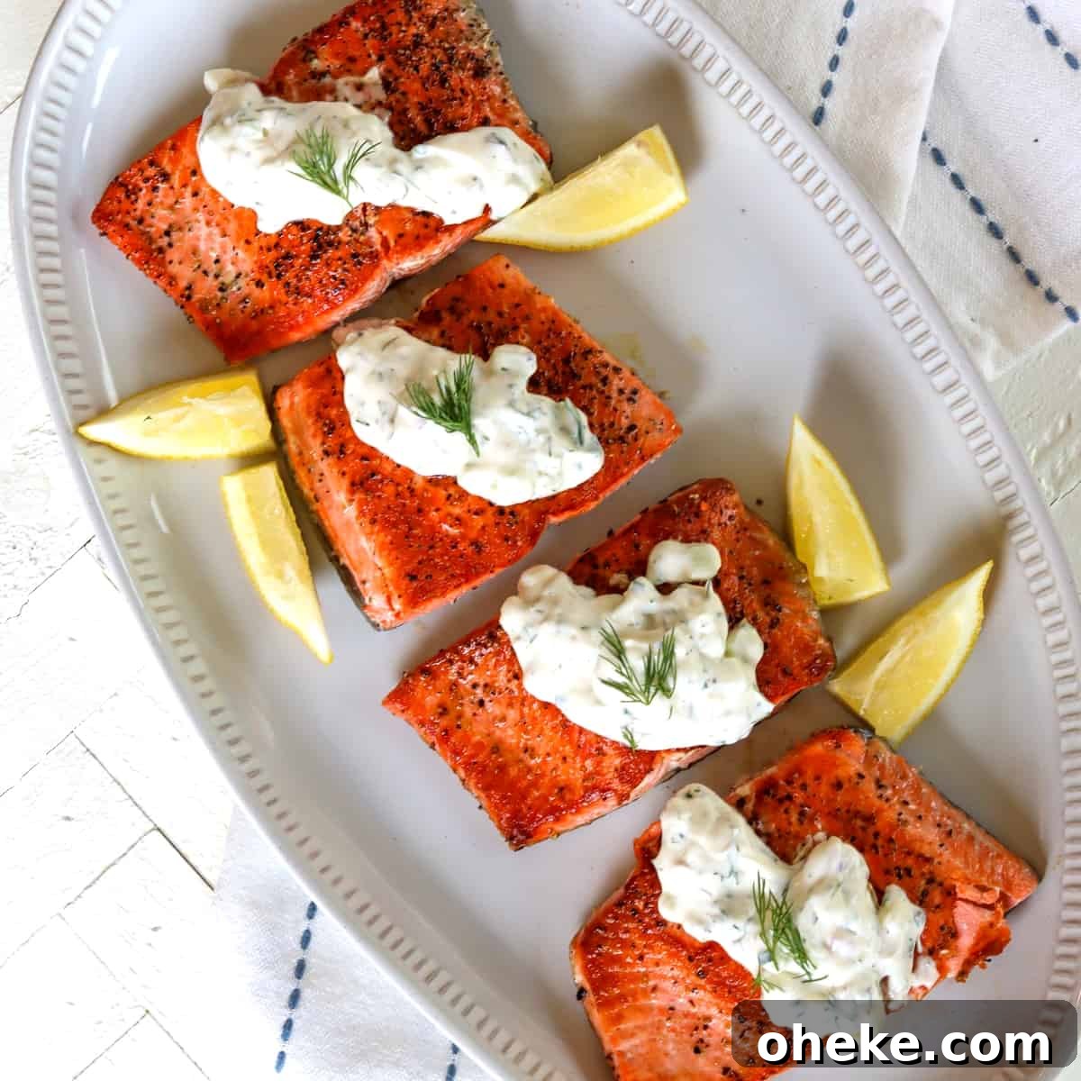 Square image of salmon with dill sauce served on an oval platter with lemon wedges.