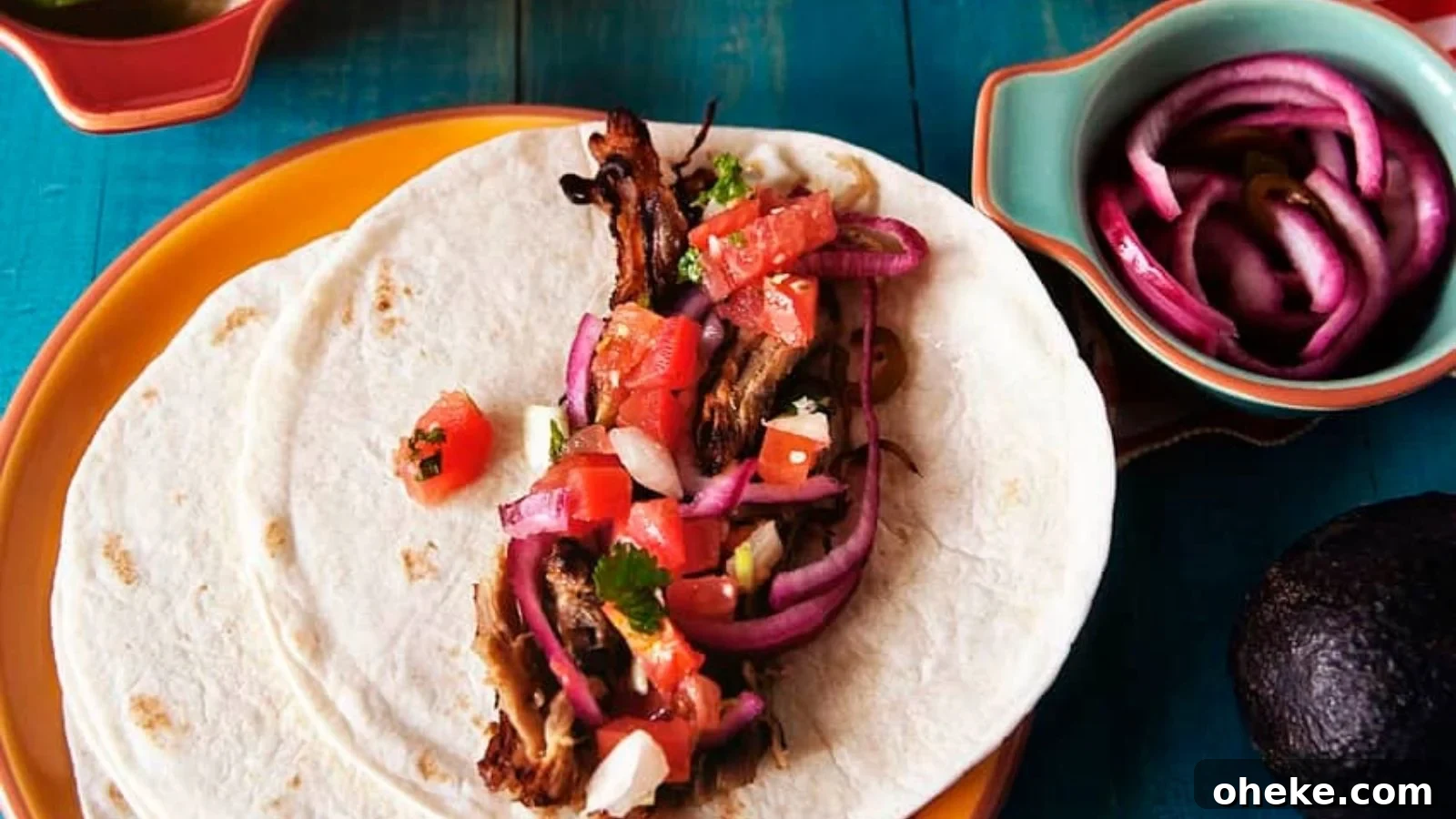 Two tortillas with meat, chopped tomatoes, onions, and herbs, on an orange plate. A bowl with sliced onions and an avocado are nearby on a blue table.
