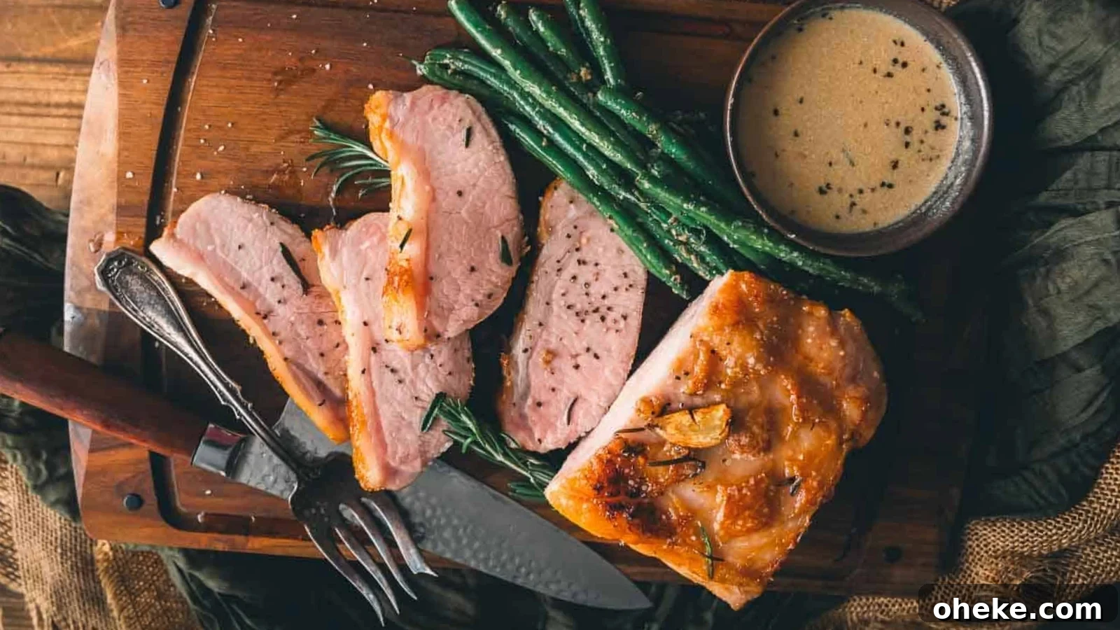 Sliced roasted pork with green beans, a fork and knife, and a bowl of creamy sauce on a wooden cutting board.