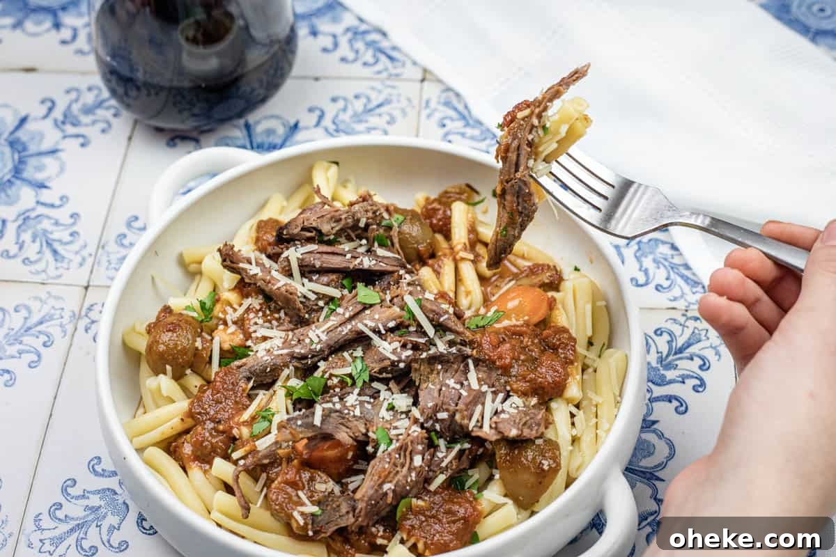 A hand lifting a fork with a bite of Italian Pot Roast Pasta on a bowl.