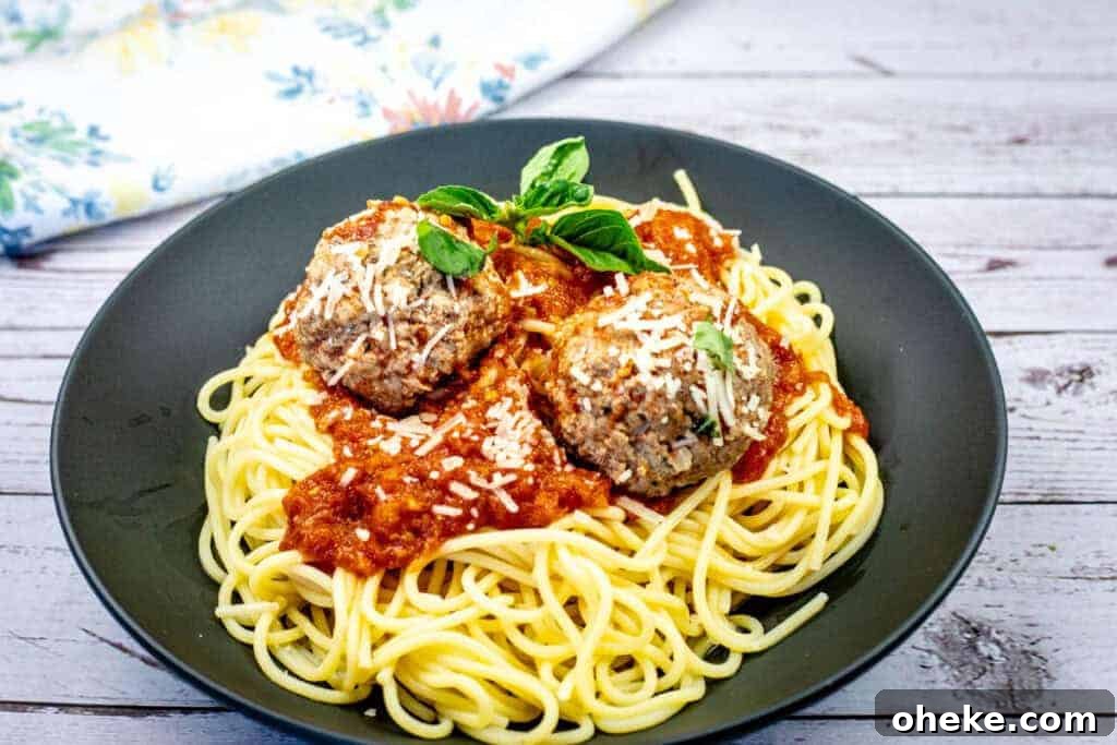 A plate of spaghetti topped with two meatballs, marinara sauce, grated cheese, and basil garnish.